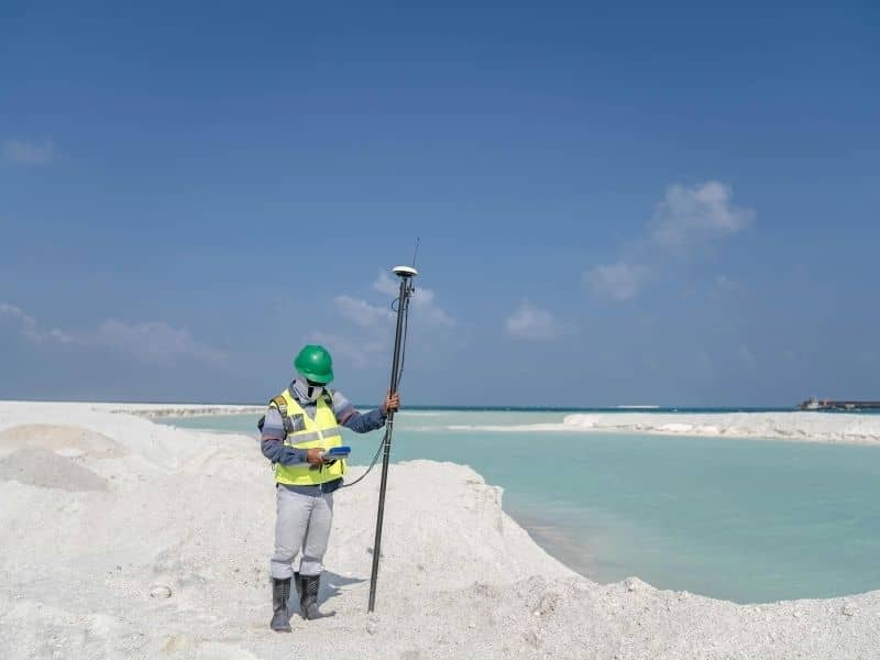 Surveyor using GNSS equipment to measure shoreline conditions and document coastal boundary changes along a waterfront property.