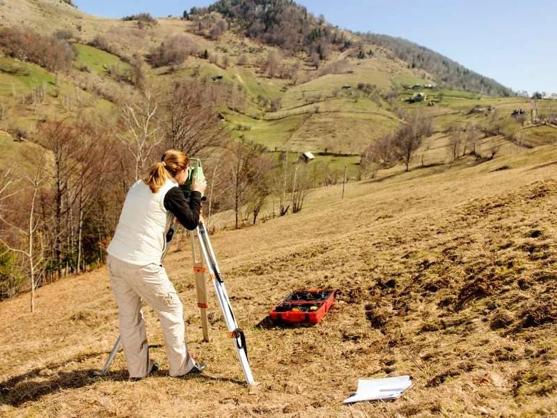Surveyor using field equipment on steep hillside terrain to measure elevation changes for topographic survey and site planning.